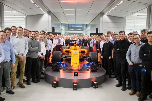 Gruppo di ingegneri e tecnici McLaren posa in fabbrica dietro una vettura da Formula 1, riflettendo le opportunità di lavoro in McLaren